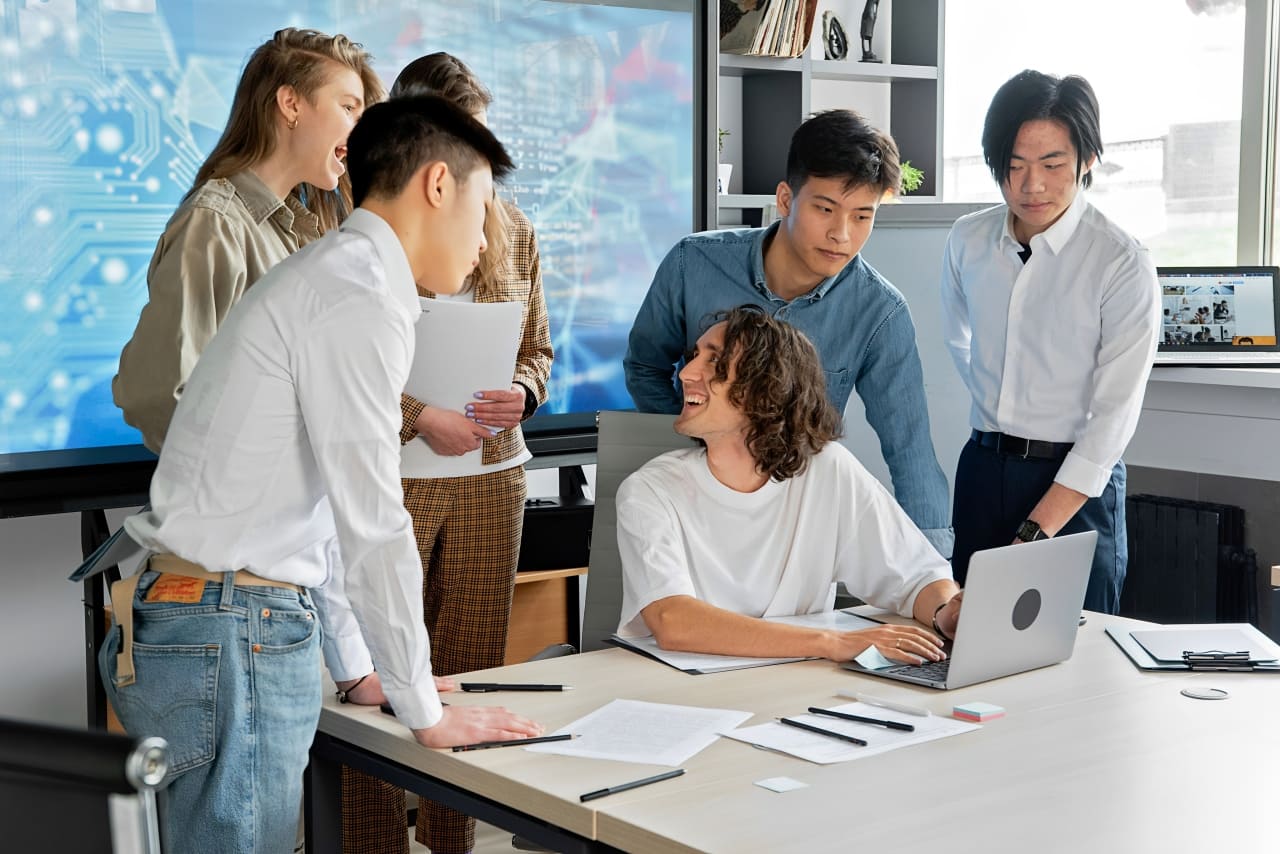 Grupo de jóvenes en una oficina colaborando con tecnología, representando nuestra empresa como especialista en capacitación en Tecnologías de la Información (TI)